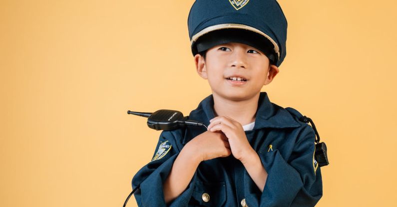 Goal Setting, Career - Pleasant Asian boy in police uniform and cap looking away while standing with hands near chest in studio on yellow background