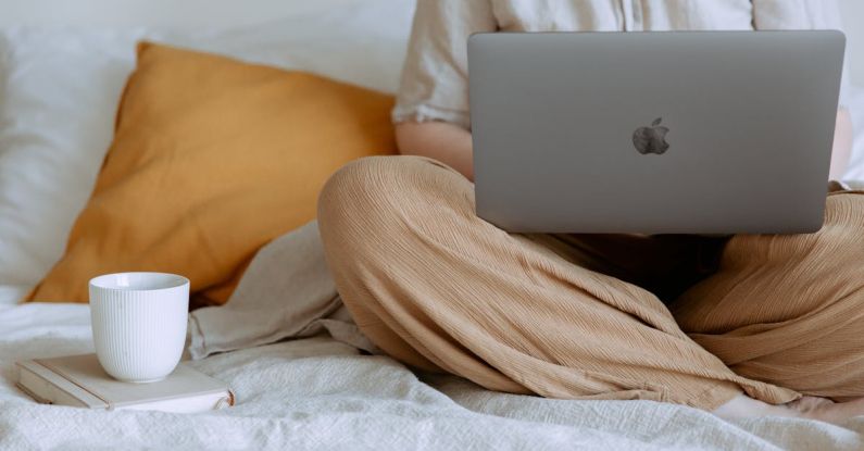 Creativity, Career - Crop faceless female in casual outfit sitting on bed with legs crossed holding laptop on knees with cup of coffee standing on notebook while working from home