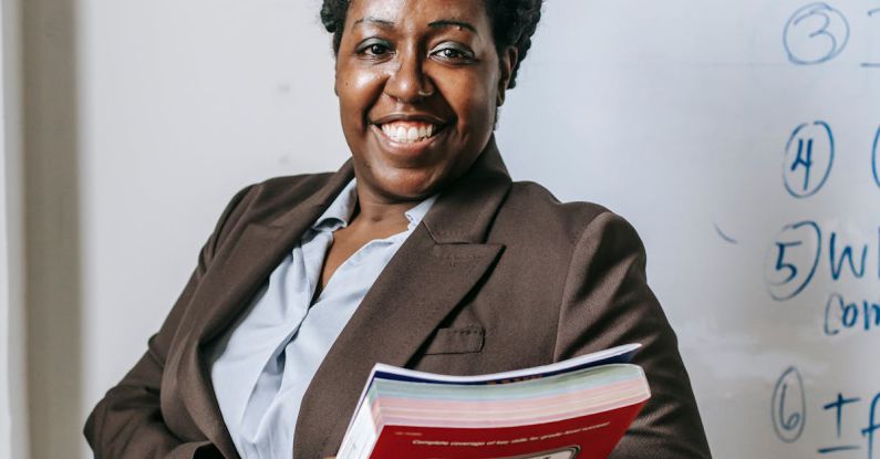 Benefits, Career Mentor - Happy black female teacher standing with workbooks near whiteboard