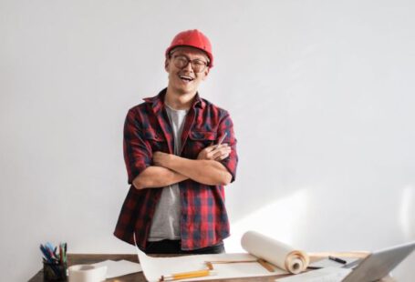 Professional Development, Career Advancement - Smiling casual man in hardhat and glasses holding arms crossed looking at camera while standing at desk with paper draft and stationery