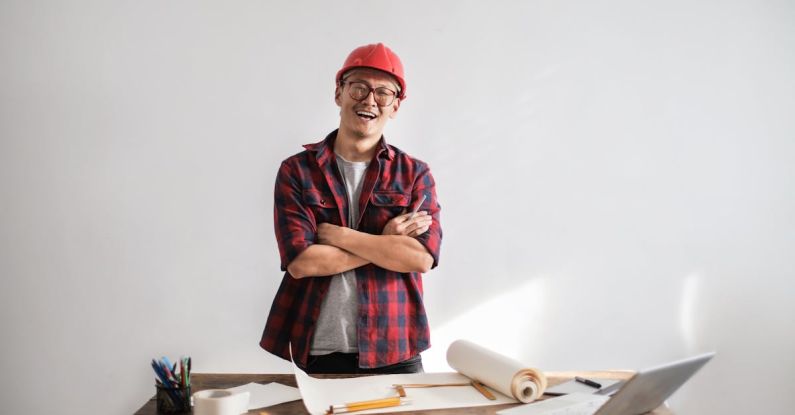 Professional Development, Career Advancement - Smiling casual man in hardhat and glasses holding arms crossed looking at camera while standing at desk with paper draft and stationery