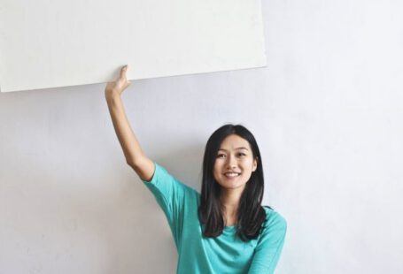 Innovations, Real Estate - Cheerful Asian woman sitting cross legged on floor against white wall in empty apartment and showing white blank banner