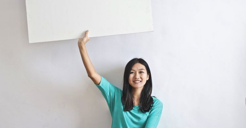 Innovations, Real Estate - Cheerful Asian woman sitting cross legged on floor against white wall in empty apartment and showing white blank banner